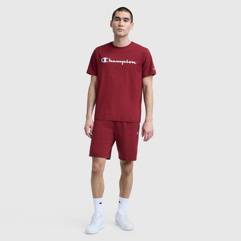 A young man stands against a plain background wearing Champion's 9" Lightweight Lounge Shorts with the C Patch Logo and a matching maroon t-shirt featuring bold Champion lettering, paired with white crew socks and sneakers for a comfortable lounge look.