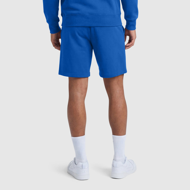 A person faces away from the camera, wearing a blue sweatshirt and Champion 9" Lightweight Lounge Shorts with C Patch Logo, white socks, and white sneakers against a plain light background.