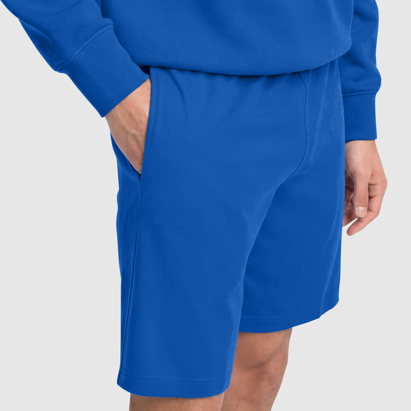 A person is shown from waist to knees wearing Champion 9" Lightweight Lounge Shorts with C Patch Logo in blue cotton jersey, one hand in their pocket, set against a plain light gray background.