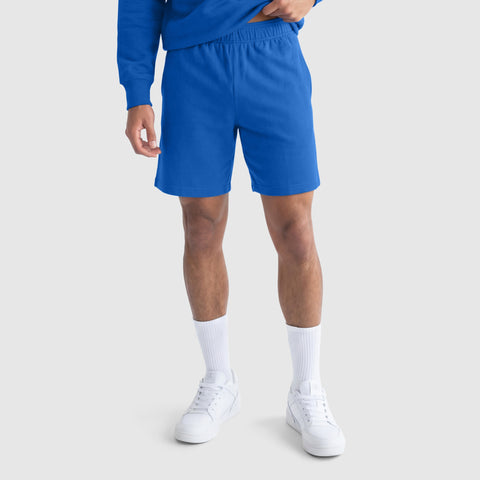 A person wears Champion’s 9" Lightweight Lounge Shorts with a C Patch Logo, a blue sweatshirt, white crew socks, and white sneakers, standing against a plain light-gray background. The upper part of the face is not visible.