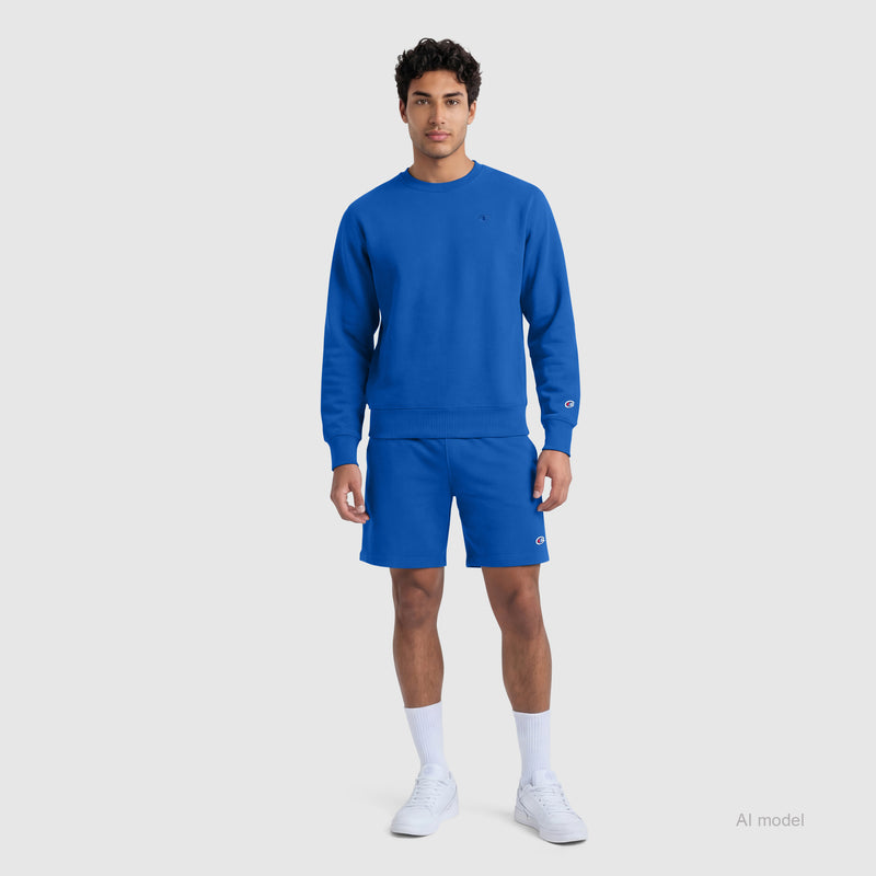 A man stands facing forward, wearing Champion's 9" Lightweight Lounge Shorts with the C Patch Logo, a matching bright blue sweatshirt, white socks, and white sneakers against a plain light gray background.