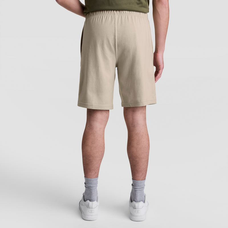 A person faces away from the camera, wearing beige Champion 9" Lightweight Lounge Shorts with a C Patch Logo, white socks, white sneakers, and an olive green shirt against a plain light background.