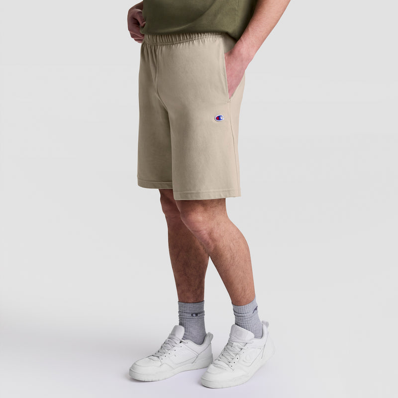A person wearing Champion 9" Lightweight Lounge Shorts, C Patch Logo in beige, a green shirt, gray socks, and white sneakers stands with hands in pockets against a plain white background.