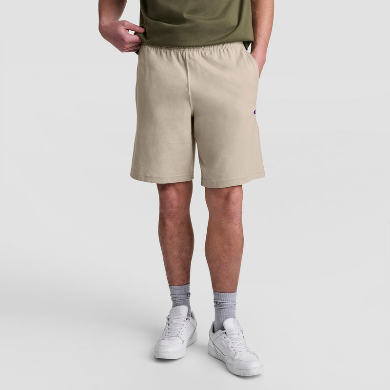 A person in a green shirt, gray socks, white sneakers, and Champion 9" Lightweight Lounge Shorts with C Patch Logo stands with one hand in their pocket against a plain white background.