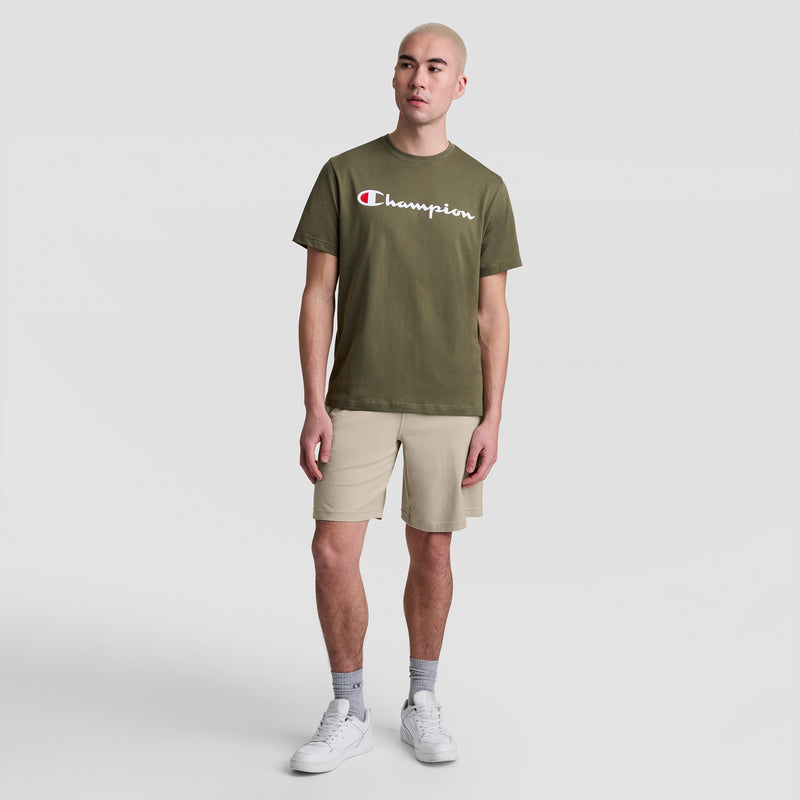 A person stands against a plain white background wearing an olive green Champion t-shirt, gray socks, white sneakers, and beige Champion 9" Lightweight Lounge Shorts with a C Patch Logo.