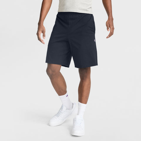 A person wearing a light gray t-shirt, Champion 9" Lightweight Lounge Shorts with the C Patch Logo in black cotton jersey, white socks, and white sneakers stands against a plain light background. Only the body from the shoulders down is visible.