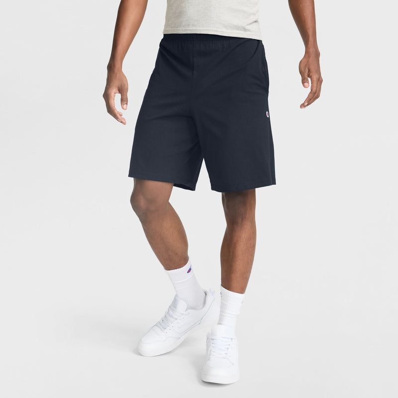 A person wearing a light gray t-shirt, Champion 9" Lightweight Lounge Shorts with the C Patch Logo in black cotton jersey, white socks, and white sneakers stands against a plain light background. Only the body from the shoulders down is visible.