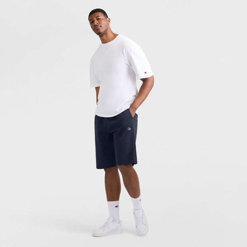 A man stands against a plain white background, wearing a white t-shirt, Champion 9" Lightweight Lounge Shorts with C Patch Logo (Big & Tall) in navy, white socks, and white sneakers. He has one hand in his pocket and looks slightly upward with a relaxed expression.