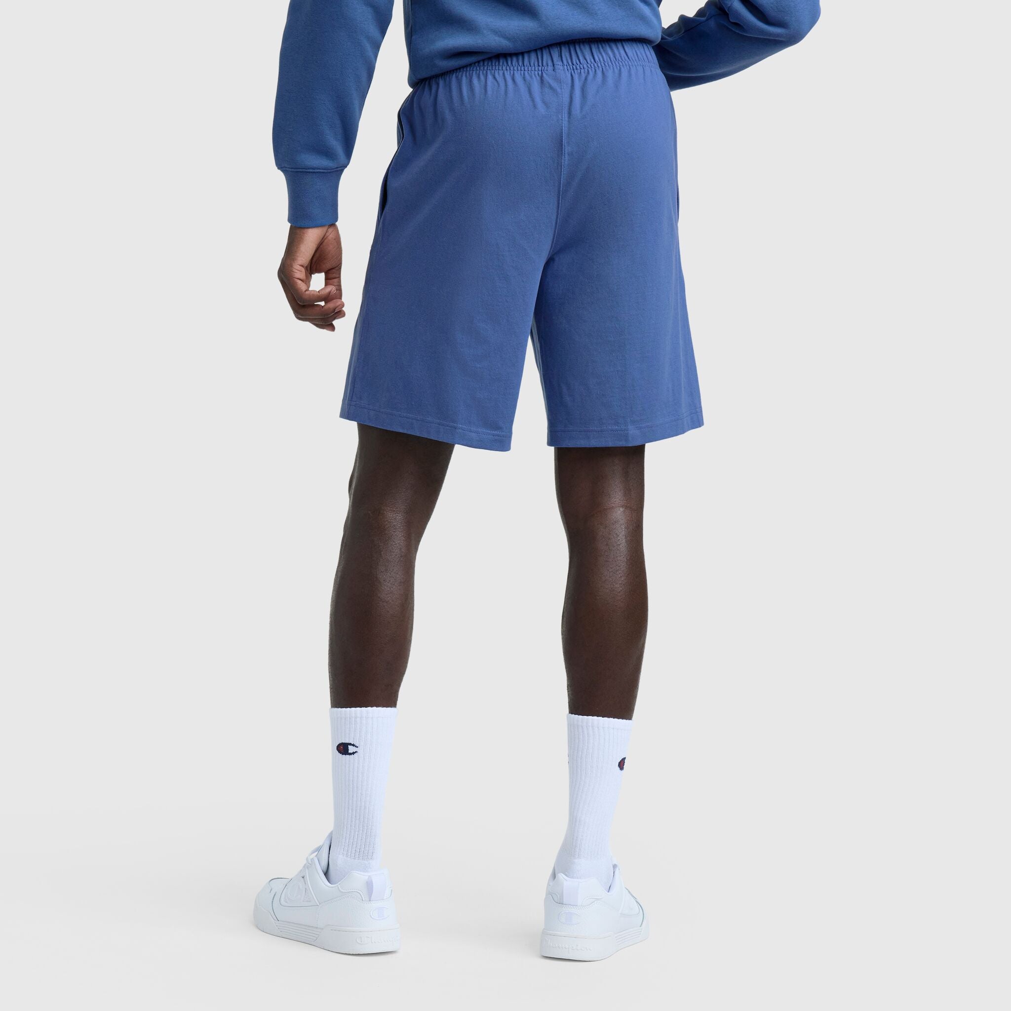 A person faces away in a blue sweatshirt and Champion 9" Lightweight Lounge Shorts with C Patch Logo, white crew socks with a logo, and white sneakers, against a plain light background.