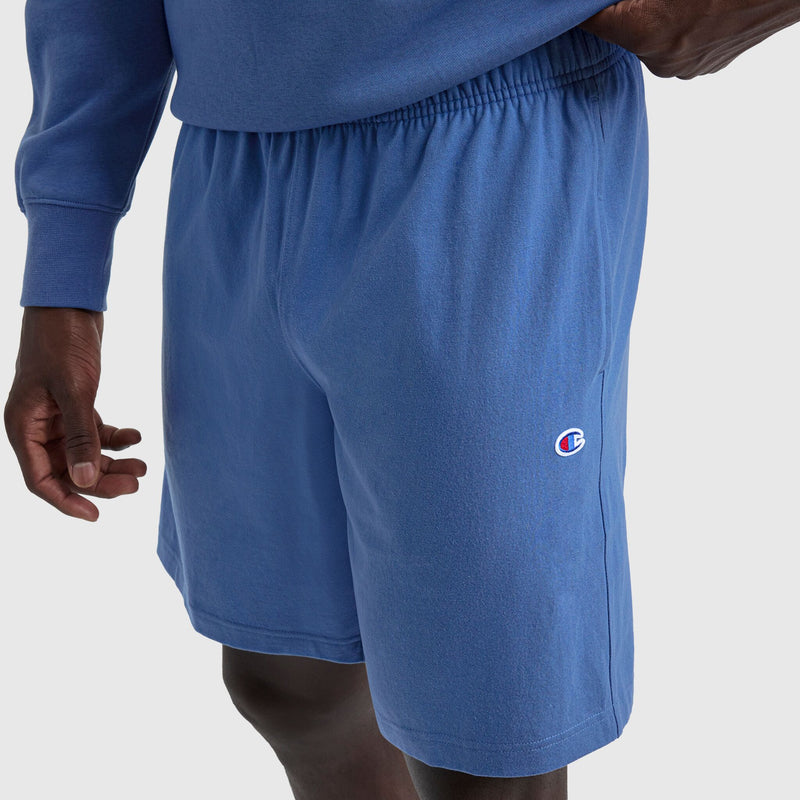 A person wearing blue Champion 9" Lightweight Lounge Shorts with a C Patch Logo and an adjustable fit, paired with a matching blue long-sleeve shirt, stands against a plain light background, one hand resting by their side.