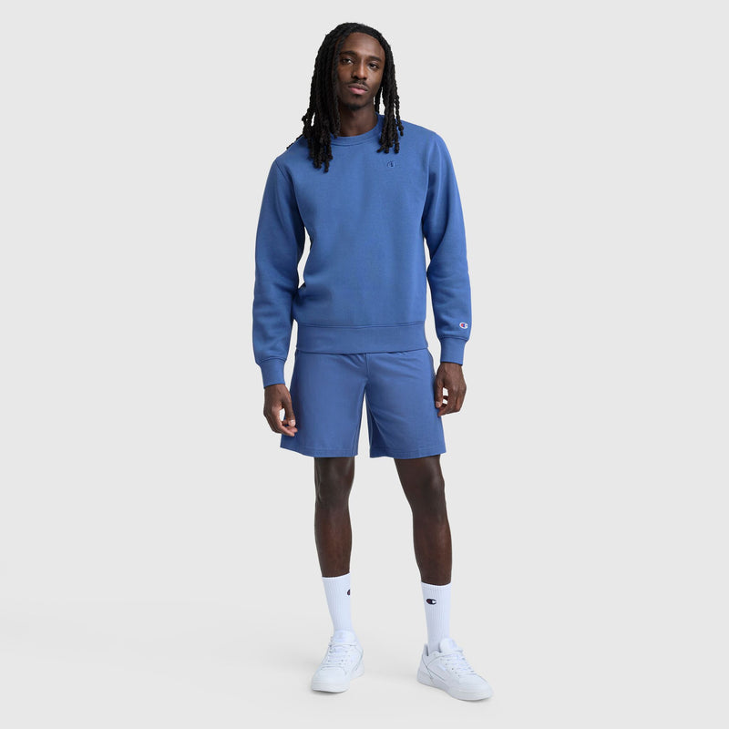A man with long dreadlocks wears Champion’s 9" Lightweight Lounge Shorts with a C Patch Logo and a matching blue sweatshirt, paired with white crew socks and sneakers, standing against a plain light gray background.