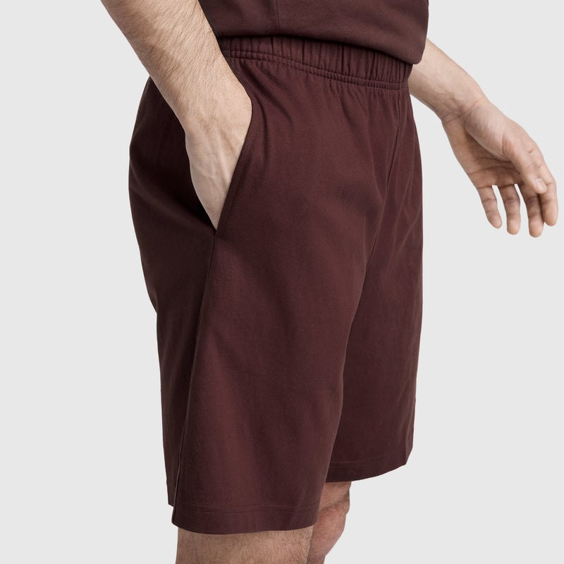 A person wearing Champion’s 9″ Lightweight Lounge Shorts with C Patch Logo in maroon, paired with a matching shirt, stands with one hand in their pocket against a light gray background.
