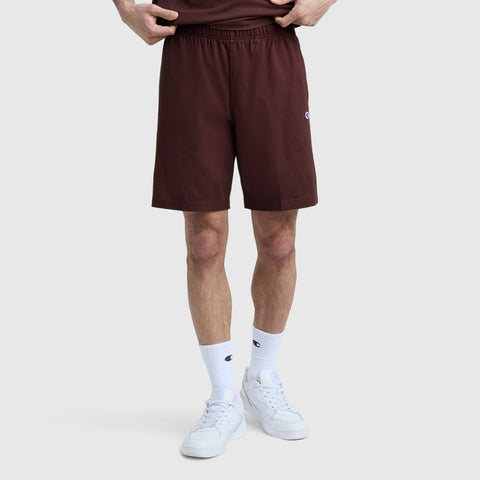 A person wearing Champion 9″ Lightweight Lounge Shorts with the C Patch Logo in maroon, paired with a matching maroon shirt, white crew socks, and white sneakers stands against a light gray background. Only the lower half of the body is visible.