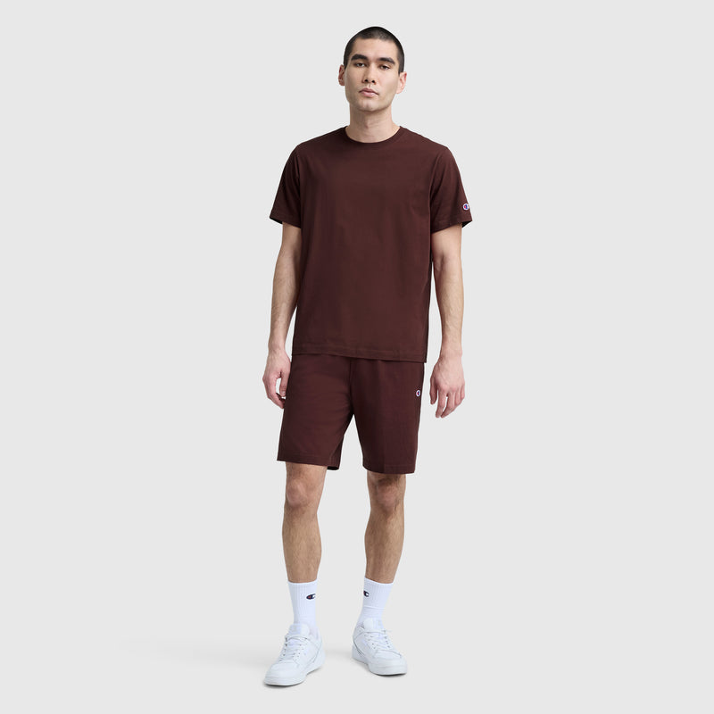 A man stands against a light gray background wearing matching burgundy Champion 9″ Lightweight Lounge Shorts with a C Patch Logo and a coordinating T-shirt, paired with white crew socks and white sneakers. He has short dark hair and a neutral expression.