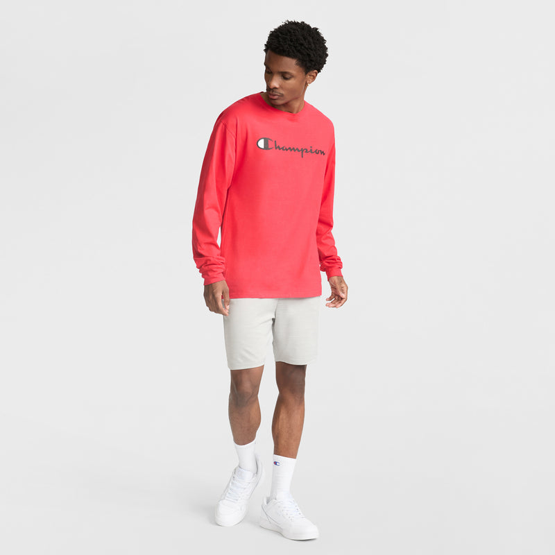 A man in a bright red Champion long-sleeve shirt, Champion 7" Moisture Wicking Sport Shorts in light gray, white socks, and white sneakers stands against a plain white background, looking down to his left.