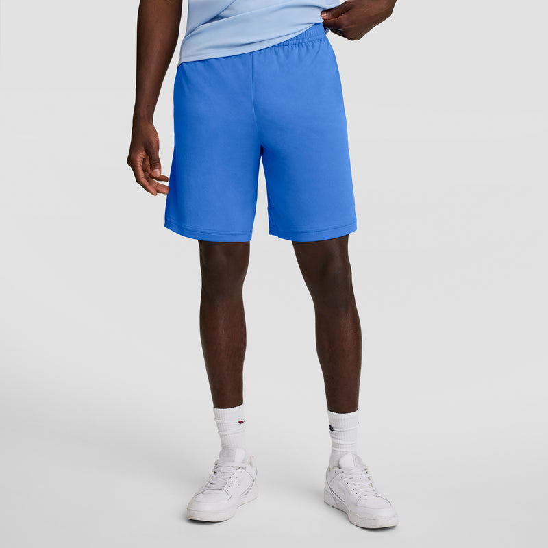 A person wearing Champion 7" Moisture Wicking Sport Shorts in bright blue, a light blue shirt, white socks, and white sneakers stands against a plain light gray background. The shorts offer all-day comfort with moisture-wicking fabric.