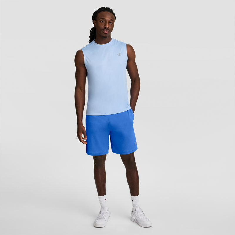 A man with long hair stands against a plain background, wearing a light blue sleeveless shirt, Champion 7" Moisture Wicking Sport Shorts in bright blue, white crew socks, and white sneakers. He looks relaxed with one hand by his side.