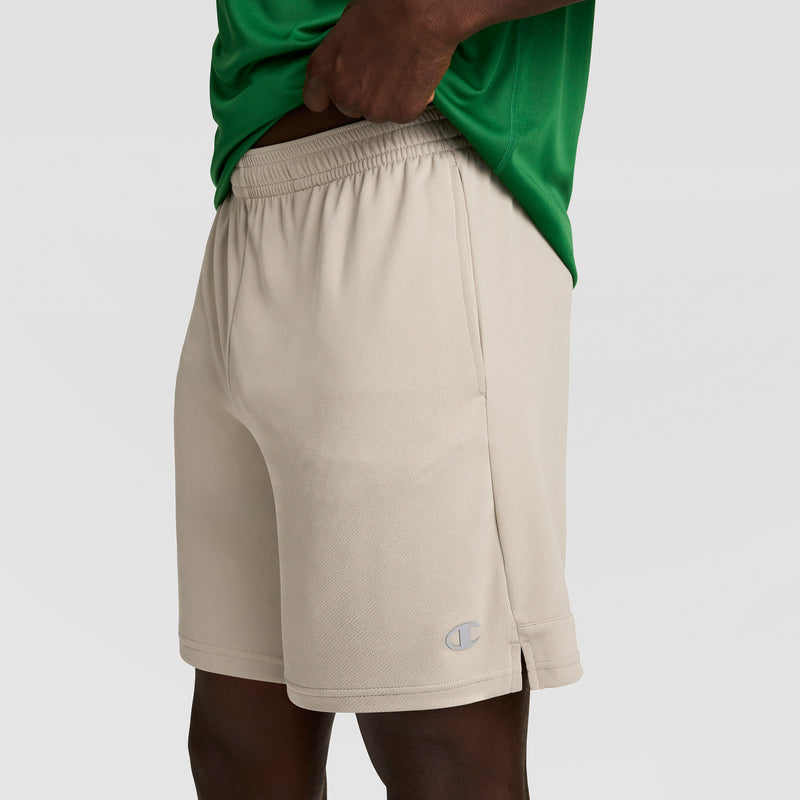 A person wears light beige Champion 7" Moisture Wicking Sport Shorts and a green shirt, with one hand lifting the shirt at the waist. The Champion logo is near the hem of these men's sport shorts featuring DOUBLE DRY technology.