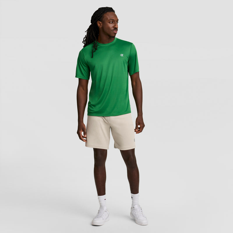 A man stands against a plain background wearing a green short-sleeve shirt and Champion 7" Moisture Wicking Sport Shorts for men with DOUBLE DRY tech, paired with white socks and sneakers. He looks left, one hand relaxed at his side.