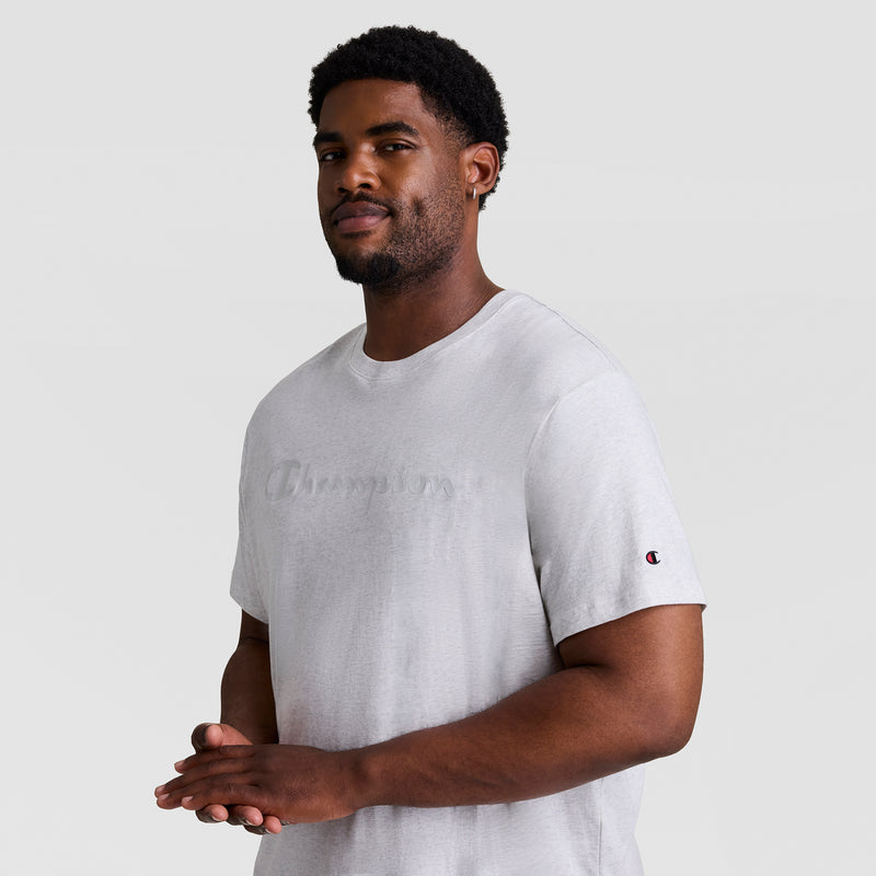 A man with short curly hair and a beard stands against a plain light background, wearing a light gray Champion Classic Graphic T-Shirt, Tonal Script Logo - Big & Tall, with the logo subtly on the front and sleeve. He looks at the camera, smiling slightly.