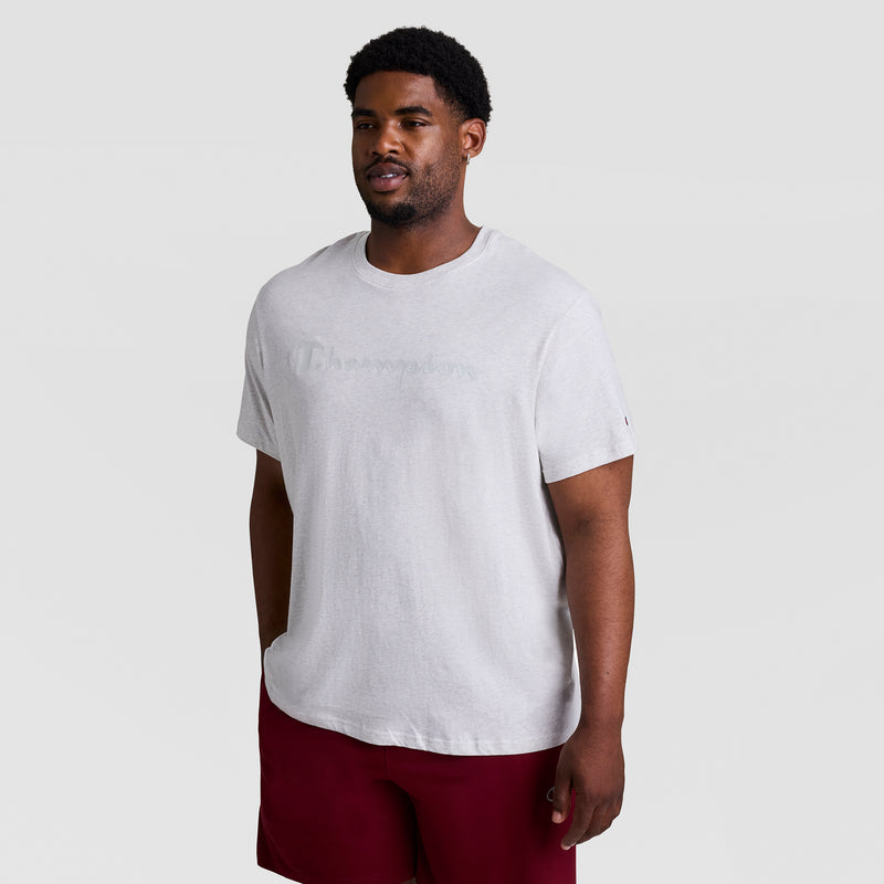 A man with short hair and a beard wears a Champion Classic Graphic T-Shirt, Tonal Script Logo - Big & Tall in light gray and red shorts, standing against a plain light gray background.
