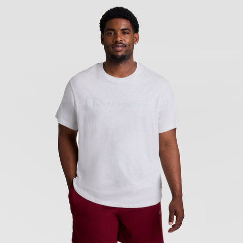 A man with short curly hair and facial hair models the Champion Classic Graphic T-Shirt, Tonal Script Logo - Big & Tall, paired with red shorts, standing against a plain light gray background with one hand in his pocket.