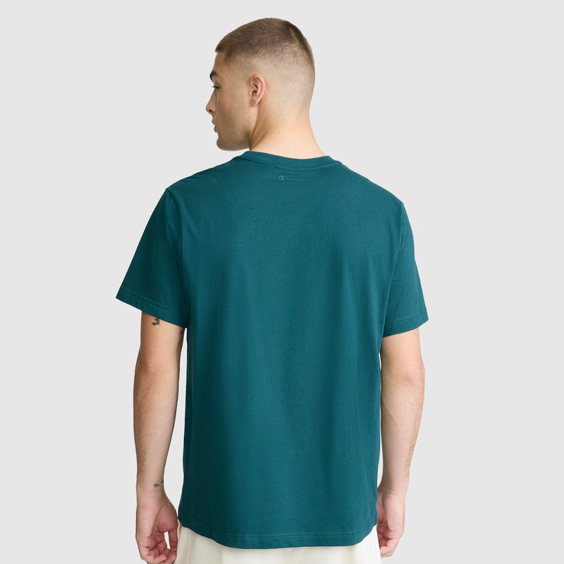 A person with short hair is standing and facing away, wearing a teal Champion Classic Graphic T-Shirt with a tonal script logo for men, made from 100% cotton, paired with light-colored pants against a plain light gray background.