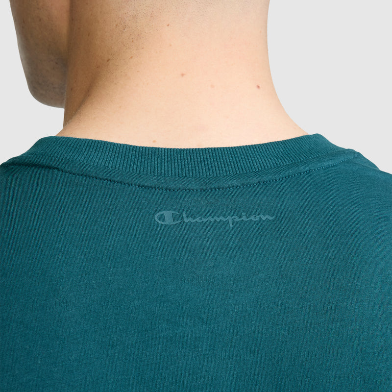 Close-up of the back of a person’s neck wearing the Champion Classic Graphic T-Shirt, Tonal Script Logo for men in teal, made from 100% cotton. The gray Champion logo is printed just below the collar, and the background is plain light gray.