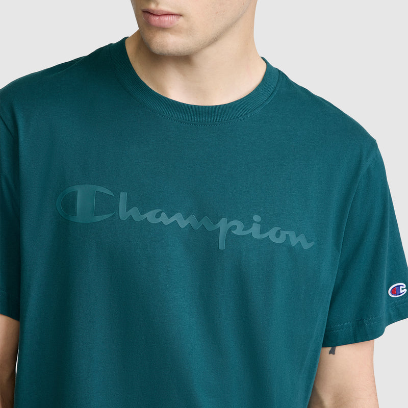 A person is wearing the Champion Classic Graphic T-Shirt for men, featuring a tonal script logo across the chest and a small logo patch on the left sleeve. The shirt is made from 100% cotton in teal, and the person’s face is partially out of frame.