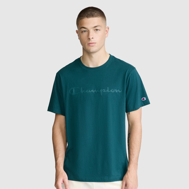 A young man wearing a teal Champion Classic Graphic T-Shirt with a tonal script logo stands against a plain light gray background, looking straight ahead with a neutral expression.