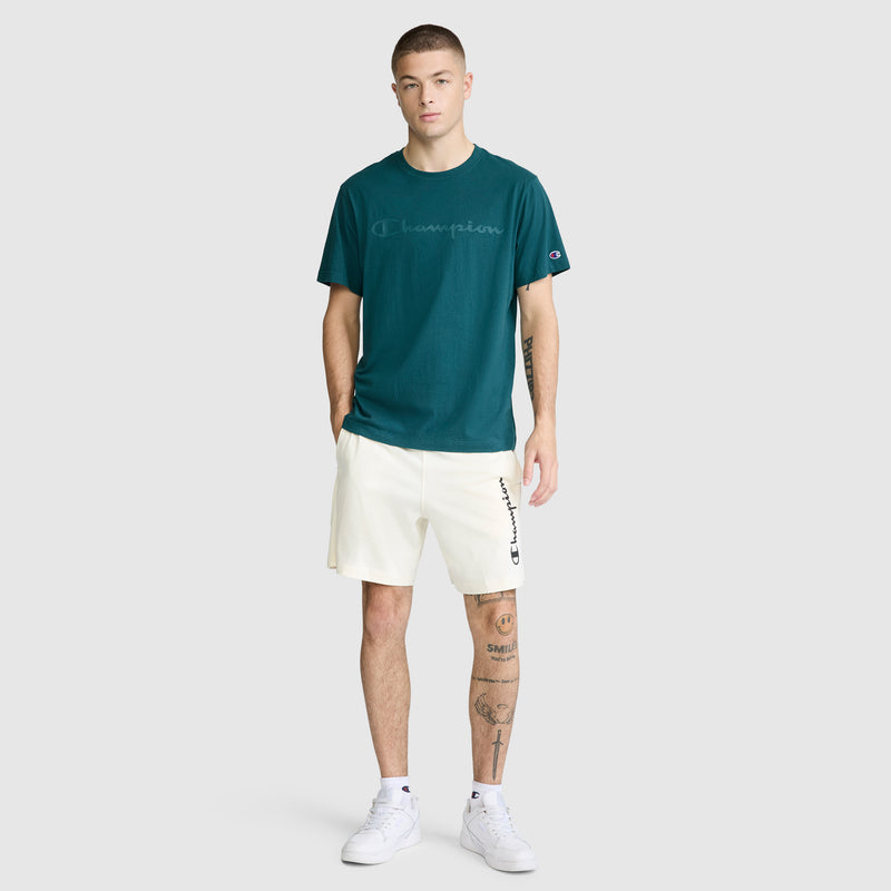 A person wearing a teal Champion Classic Graphic T-Shirt with a tonal script logo, paired with white shorts and white sneakers, stands against a plain light gray background. Their arms are relaxed by their sides, and they have visible tattoos on their left leg.