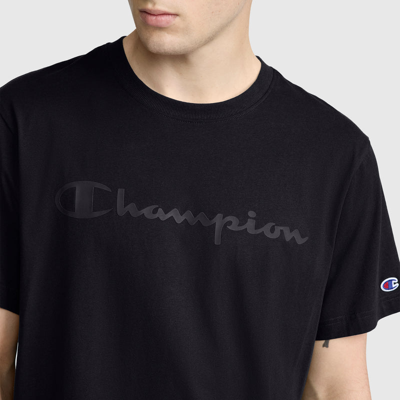 A person wearing the Champion Classic Graphic T-Shirt, featuring a tonal script logo across the chest and a small Champion patch on the left sleeve. The shirt is black and made of 100% cotton. The background is plain and light gray.
