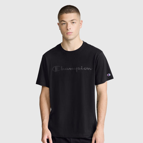 A young man wearing the Champion Classic Graphic T-Shirt with a tonal script logo stands facing forward against a plain light gray background. He has short hair and a neutral expression, highlighting the comfort of the 100% cotton fabric.