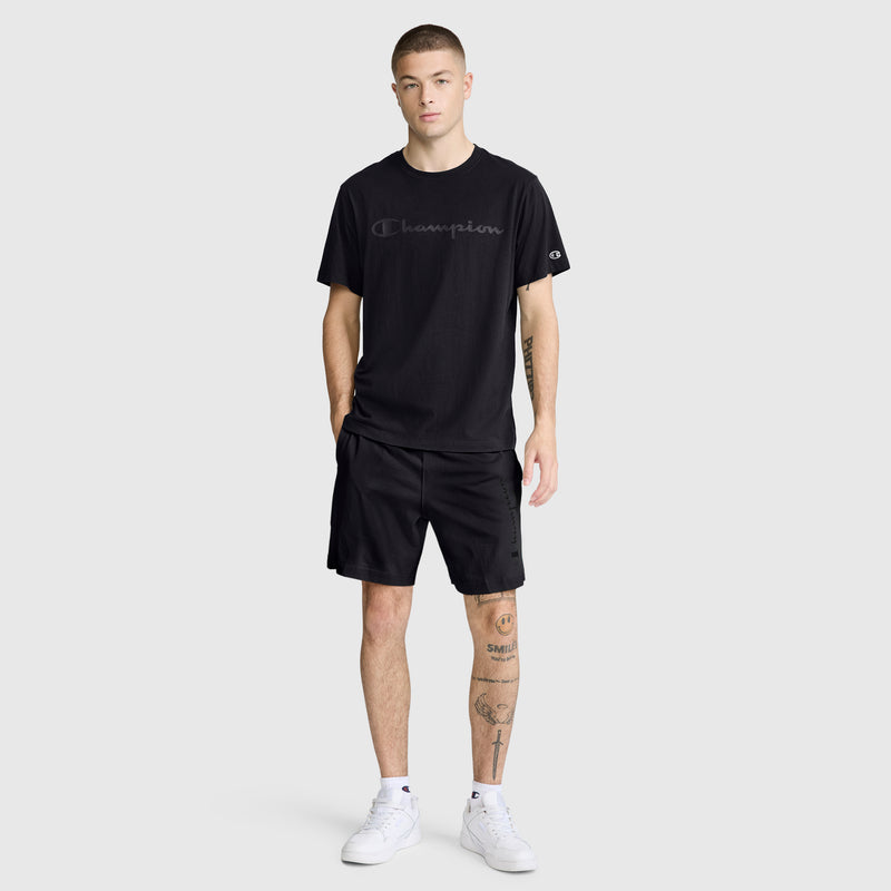 A man with short hair stands facing forward, wearing a black Champion Classic Graphic T-Shirt with a tonal script logo made from 100% cotton, paired with black shorts and white sneakers. He has tattoos on his left leg and his right hand in his pocket.