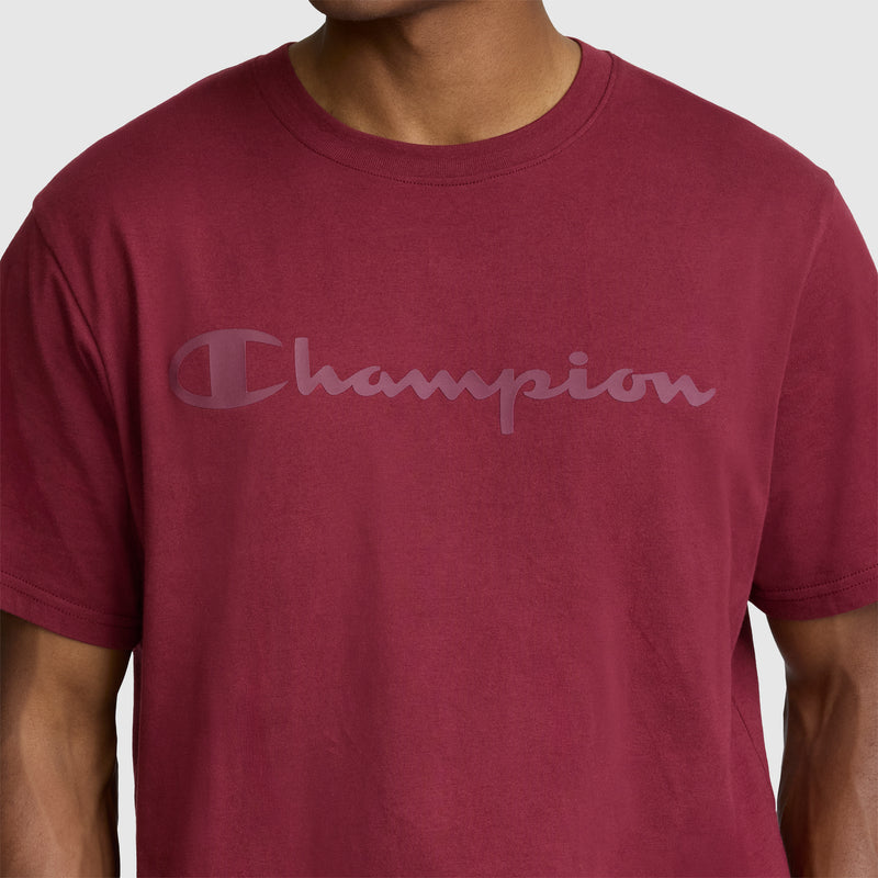 A man models the Champion Classic Graphic T-Shirt with a tonal script logo for men in maroon, made from 100% cotton, standing out against a plain, light-colored background.