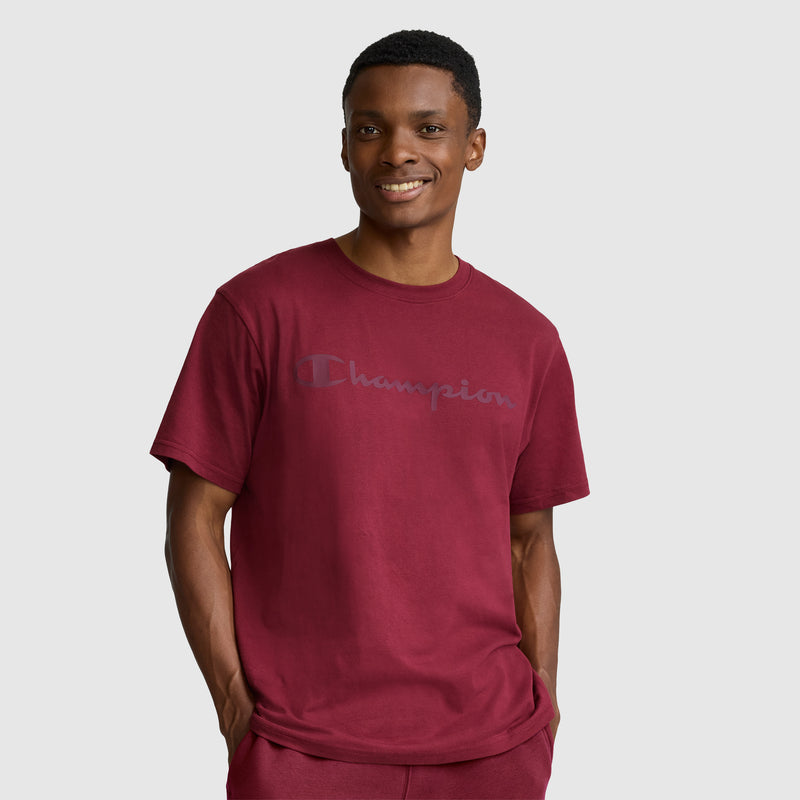 A person stands against a plain light background, smiling with their hands in their pockets while wearing the Champion Classic Graphic T-Shirt with a tonal script logo for men in maroon and matching pants. The 100% cotton fabric of the shirt ensures comfort for this casual look.