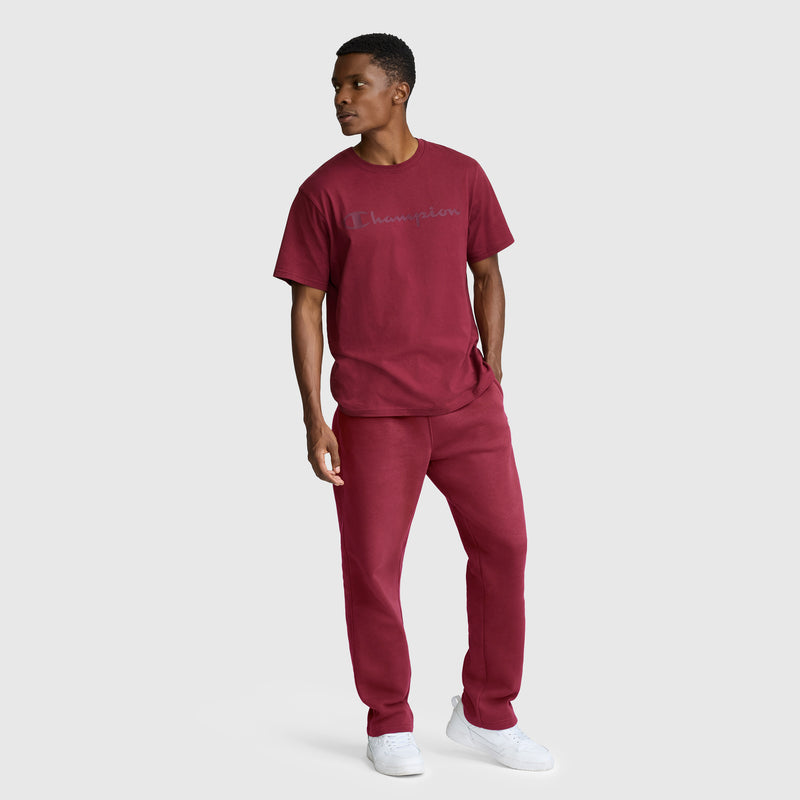 A man stands against a plain light gray background wearing a matching maroon Champion Classic Graphic T-Shirt with a Tonal Script Logo and joggers, paired with white sneakers.