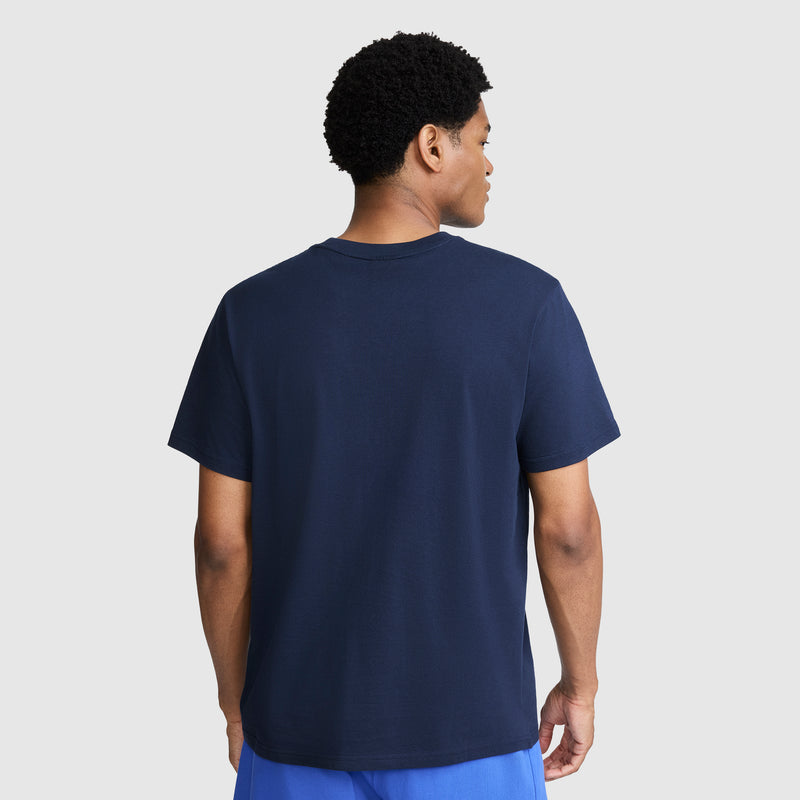 A person stands facing away, wearing a Champion Classic Graphic T-Shirt with a tonal script logo in navy blue, made from 100% cotton, paired with blue shorts, against a light gray background.