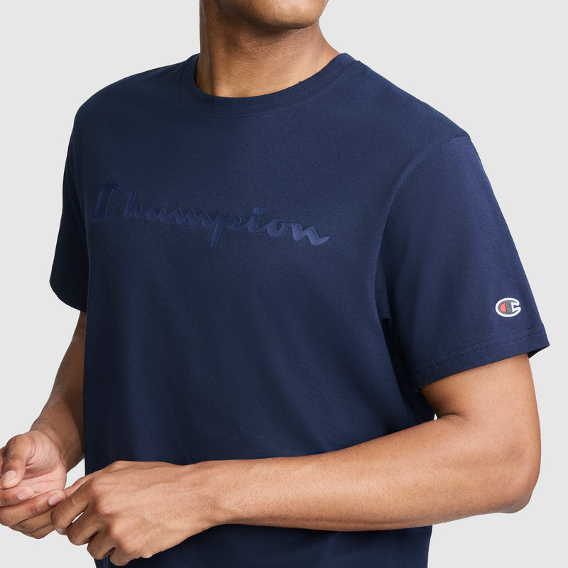 A person wearing a navy blue Champion Classic Graphic T-Shirt for men, featuring the tonal script logo on the chest and sleeve, stands against a plain light gray background.