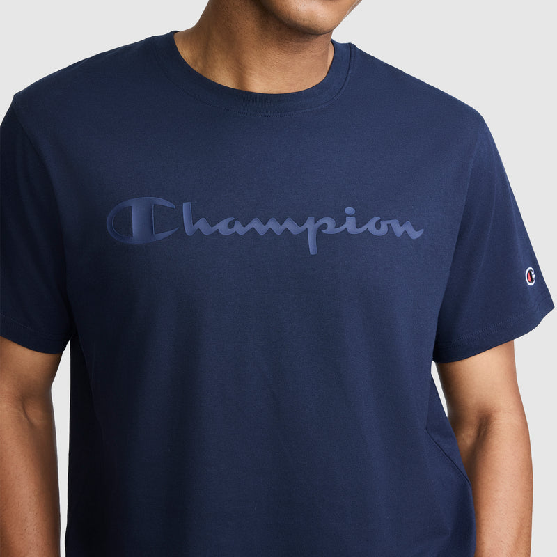 A person is wearing the Champion Classic Graphic T-Shirt, featuring a tonal script logo across the chest. The men's shirt is navy blue and made from 100% cotton. Their face is partially out of frame against a plain background.