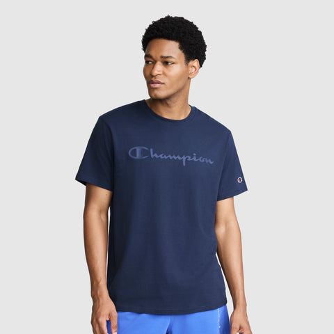A man wearing a navy blue Champion Classic Graphic T-Shirt with a tonal script logo and blue shorts stands against a plain light gray background, looking slightly to his left with a neutral expression. Made from 100% cotton, the shirt offers comfort and style for his casual look.