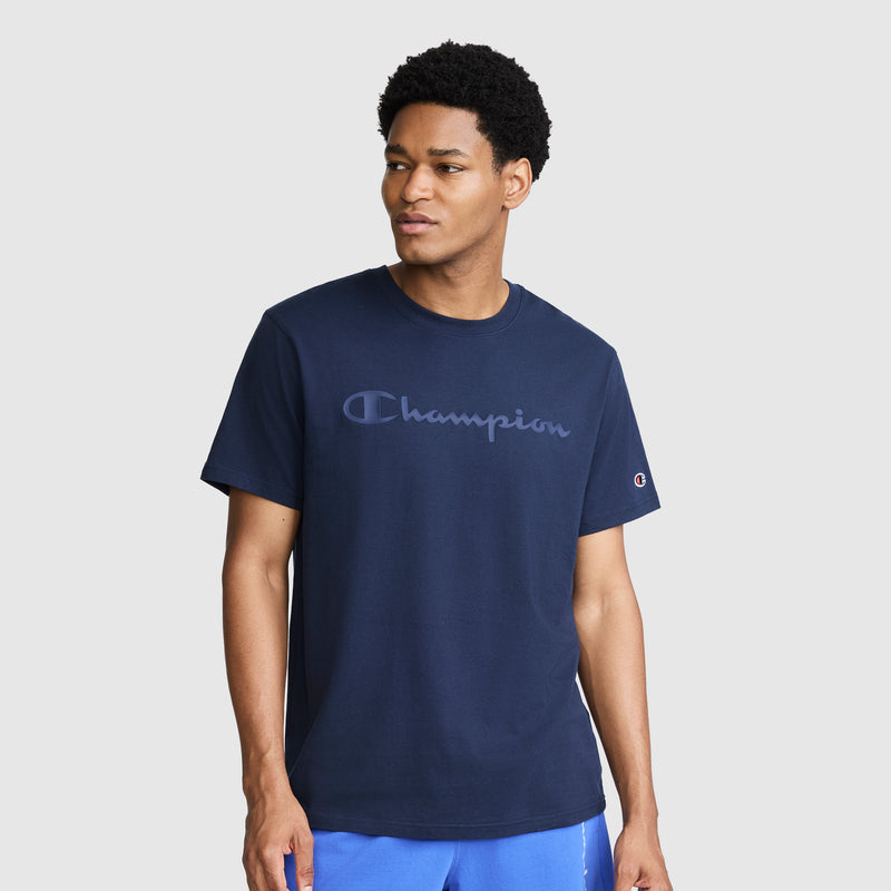 A man wearing a navy blue Champion Classic Graphic T-Shirt with a tonal script logo and blue shorts stands against a plain light gray background, looking slightly to his left with a neutral expression. Made from 100% cotton, the shirt offers comfort and style for his casual look.