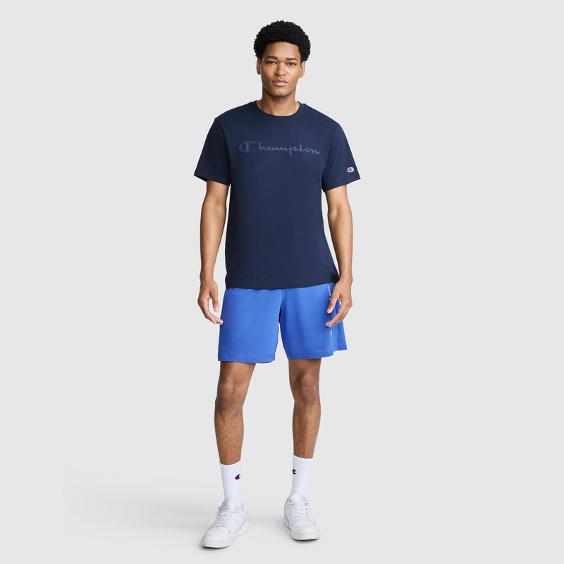 A man stands facing forward, wearing a navy blue Champion Classic Graphic T-Shirt with a tonal script logo made from 100% cotton for men, paired with bright blue shorts, white socks, and white sneakers against a plain light background.