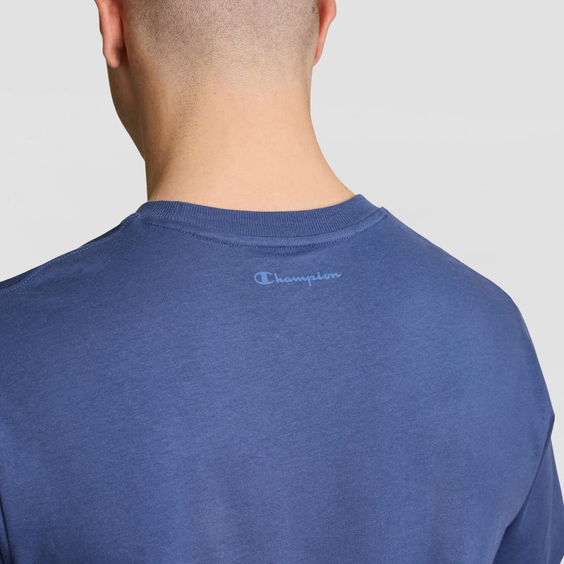 A person with a shaved head is seen from behind in a blue Champion Classic Graphic T-Shirt featuring a tonal script logo on the upper back, paired with Champion joggers for a relaxed look.