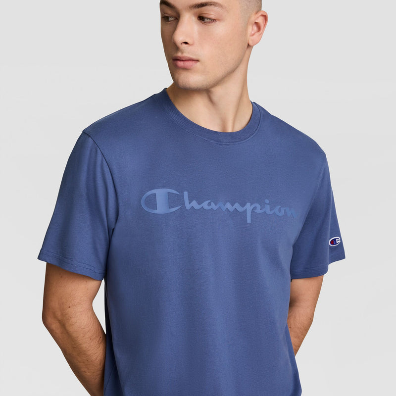A man wearing the Champion Classic Graphic T-Shirt with a tonal script logo stands with his hands behind his back, looking slightly to the side against a plain light background.