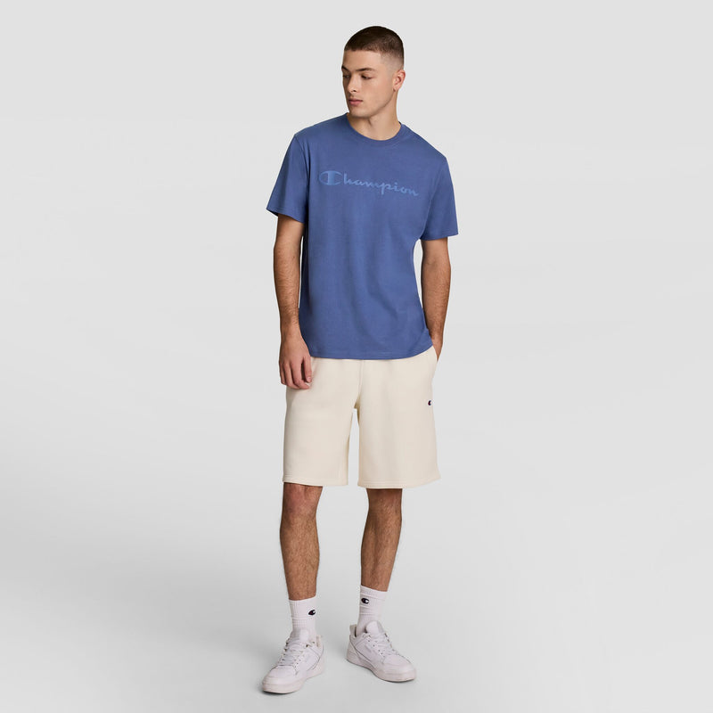 A young man stands against a plain backdrop, wearing a blue Champion Classic Graphic T-Shirt with Tonal Script Logo, cream shorts, white crew socks, and white sneakers. He looks slightly left with his hands relaxed at his sides.