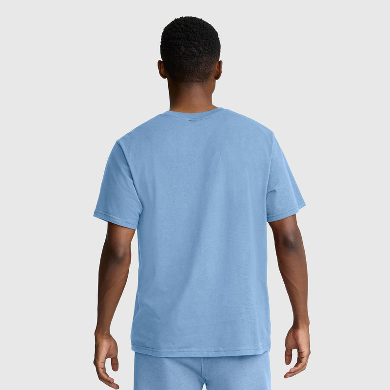 A person stands facing away from the camera, wearing a Champion Classic Graphic T-Shirt with a tonal script logo in light blue 100% cotton for men, paired with matching shorts against a plain light gray background.