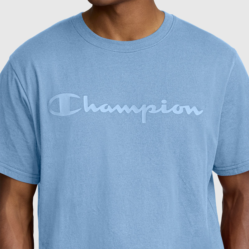 A person wearing the Champion Classic Graphic T-Shirt, Tonal Script Logo—a men’s tee made from 100% cotton in light blue, featuring “Champion” scripted across the chest in a matching blue, stylized font.