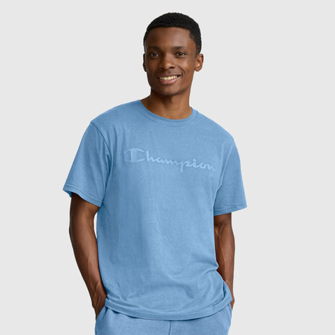 A smiling person wears the Champion Classic Graphic T-Shirt with tonal script logo in light blue, paired with matching pants, standing with hands in pockets against a plain light background.