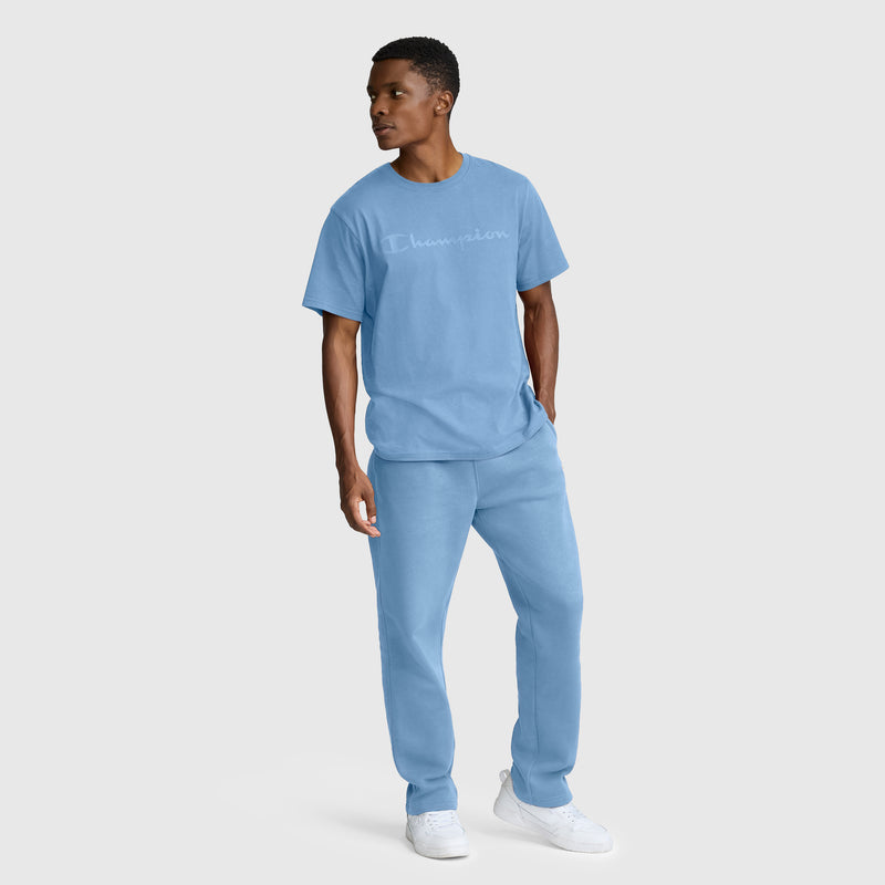 A man stands against a plain light background, wearing a light blue Champion Classic Graphic T-Shirt with a tonal script logo and matching sweatpants, paired with white sneakers. He looks to his left, his left hand in his pocket.